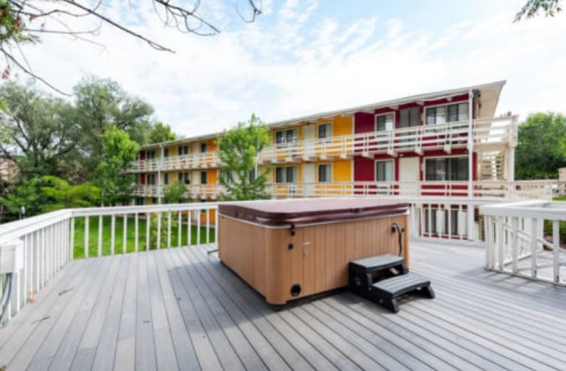 A wooden hot tub with black steps sits on a spacious gray deck, surrounded by a white railing. In the background, Buffalo Canyon Apartments Boulder rises—its three stories featuring red and yellow accents amid green trees.