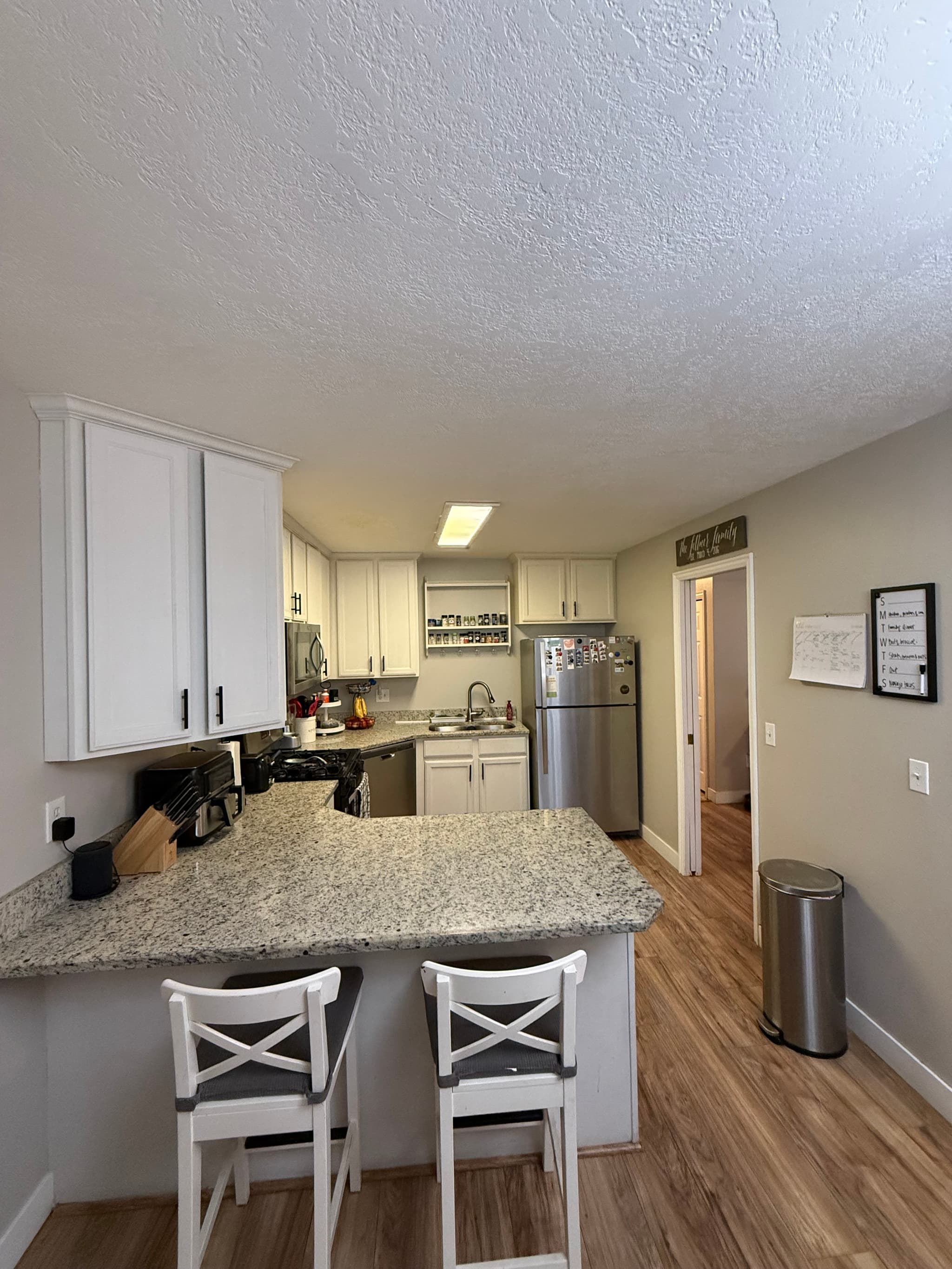 A modern kitchen at 282 W 960 S, APT C features white cabinets, stainless steel appliances, and granite countertops. Two white barstools sit at the counter, with a calendar and trash bin on the right and wood floors throughout.