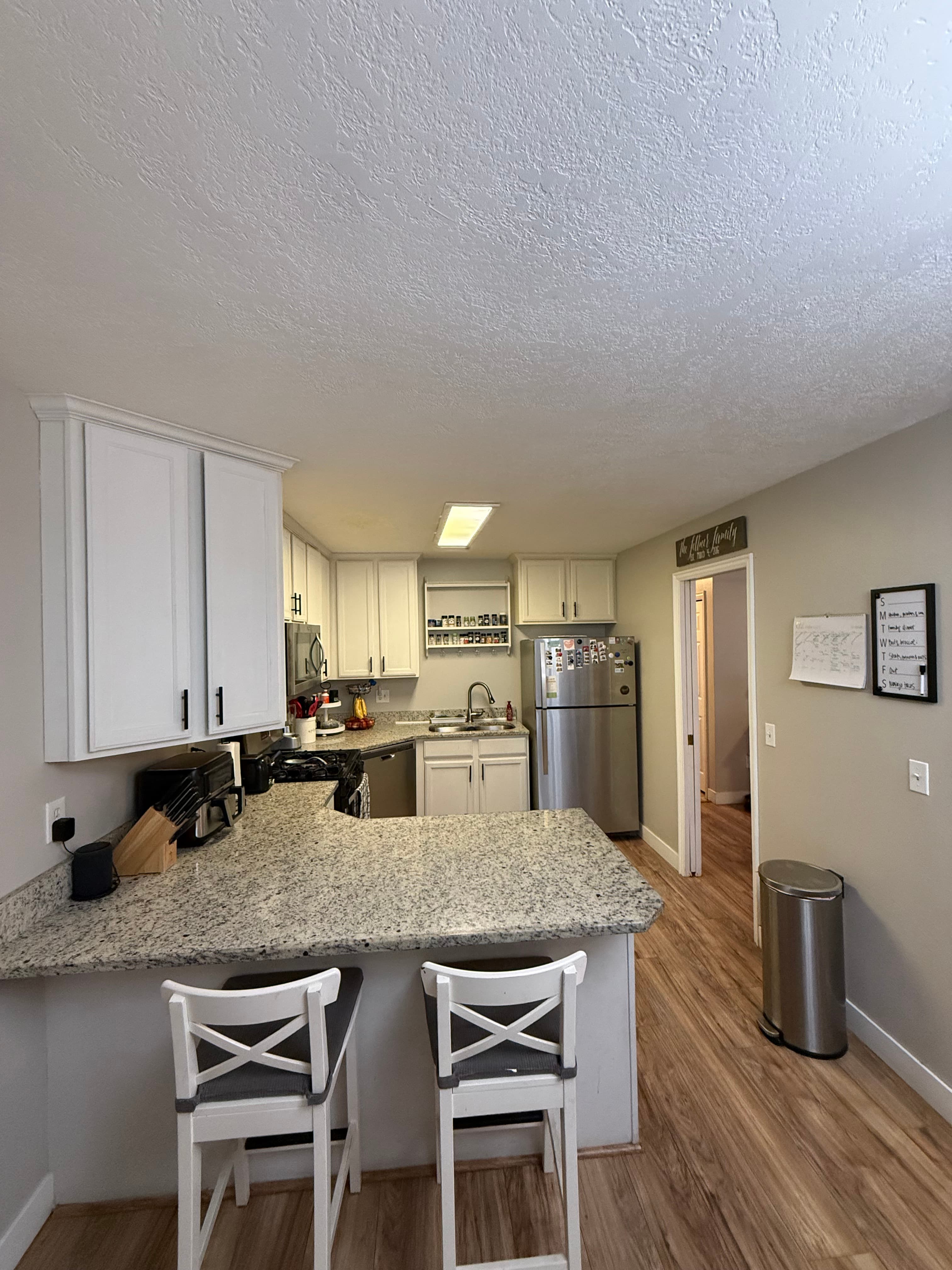 A modern kitchen at 282 W 960 S, APT C features white cabinets, stainless steel appliances, and granite countertops. Two white barstools sit at the counter, with a calendar and trash bin on the right and wood floors throughout.