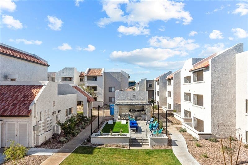 A bright, sunny courtyard at Paseo On University in Tempe, surrounded by white apartment buildings with red-tiled roofs. The student housing features a shaded lounge area with seating, a grill, and a lawn bordered by pathways and desert landscaping.
