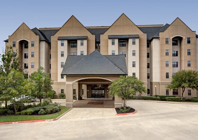 A multi-story beige hotel building, reminiscent of Callaway House student housing, features pyramid-shaped roofs, large windows, and a covered driveway entrance, all surrounded by trees and landscaped greenery under a clear blue sky.