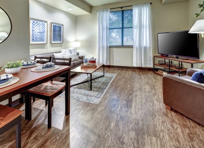 A cozy living and dining area at Callaway Villas student housing features wooden floors, a dining table with place settings, a brown sofa, coffee table, large TV, window with sheer curtains, and modern wall art—perfect for College Station living.
