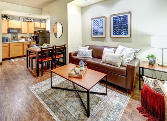 A cozy, modern apartment living area at Callaway Villas in College Station features a brown leather sofa, patterned rug, wooden coffee table, dining table with chairs, and an open kitchen—perfect student housing with neutral tones and framed art enhancing the decor.