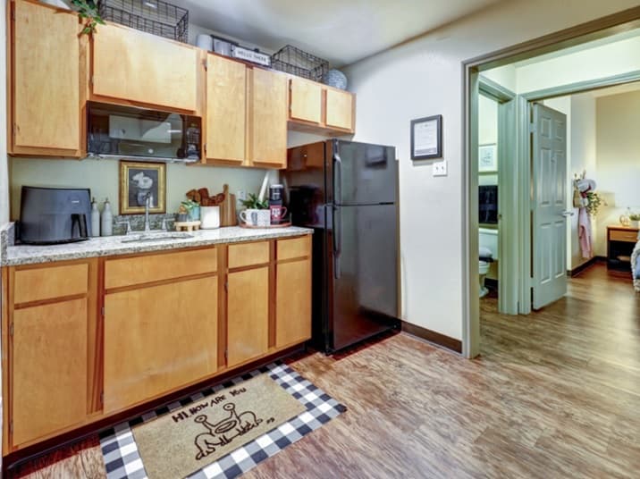 A cozy kitchen in Callaway House features wooden cabinets, granite countertop, black fridge, and small appliances. A decorative mat adorns the floor. An open door leads to a room with a bed and wooden flooring—perfect for student housing.