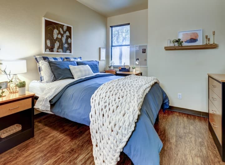 A cozy bedroom in Callaway House with a blue bedspread, plaid pillows, and a chunky white knit blanket. Nightstands with lamps, a desk by the window, wall art, and wood floors create inviting student housing with soft lighting and a tidy appearance.