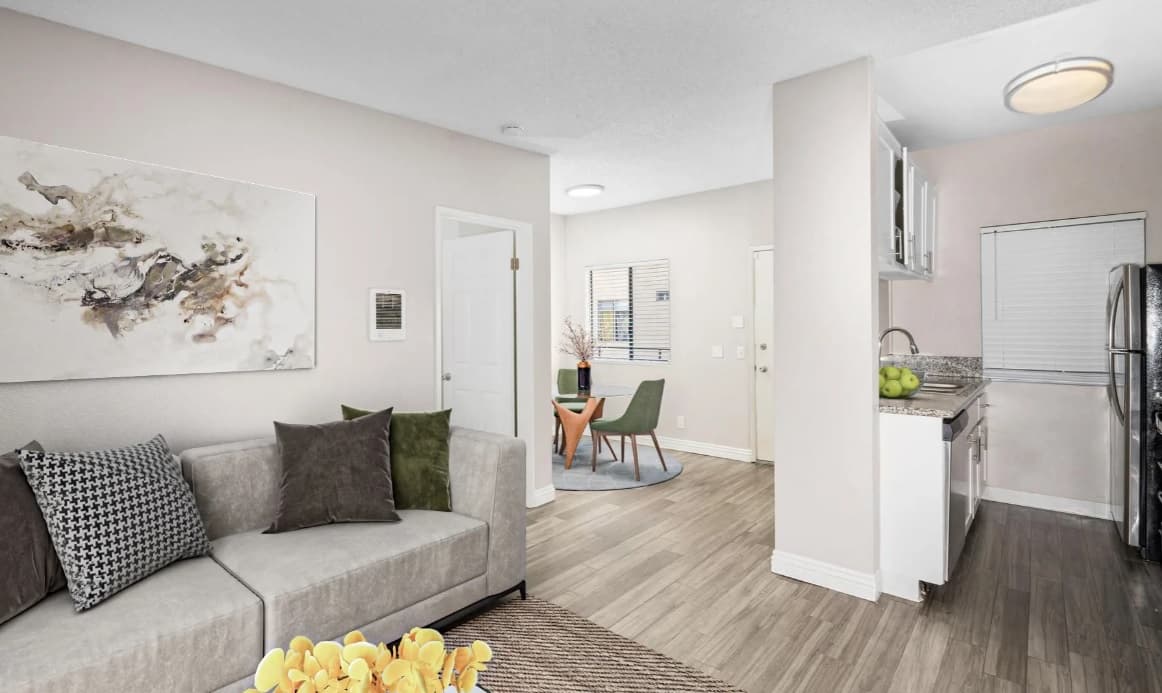Modern Campus Village Apartments interior with light wood floors, a beige sofa with assorted pillows, abstract wall art, a small dining area by the window, and a kitchen with white cabinets and stainless steel appliances.