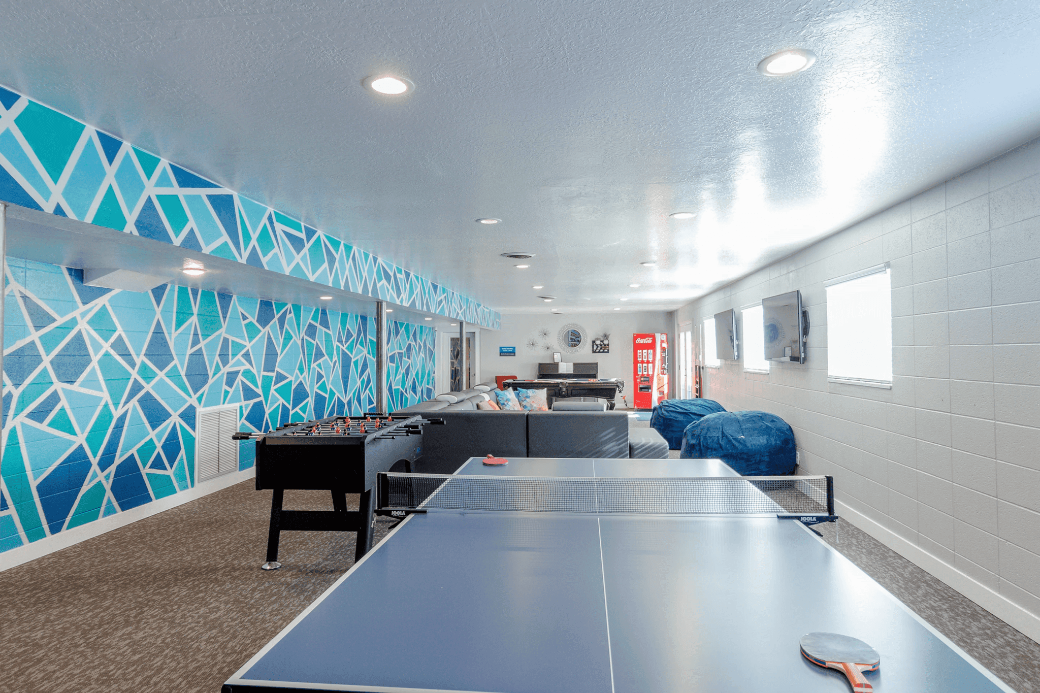 A spacious game room at Campus Way Provo features a blue geometric accent wall, foosball and ping pong tables, comfy couches, bean bags, a TV, and a red vending machine—all brightly lit with ceiling lights.