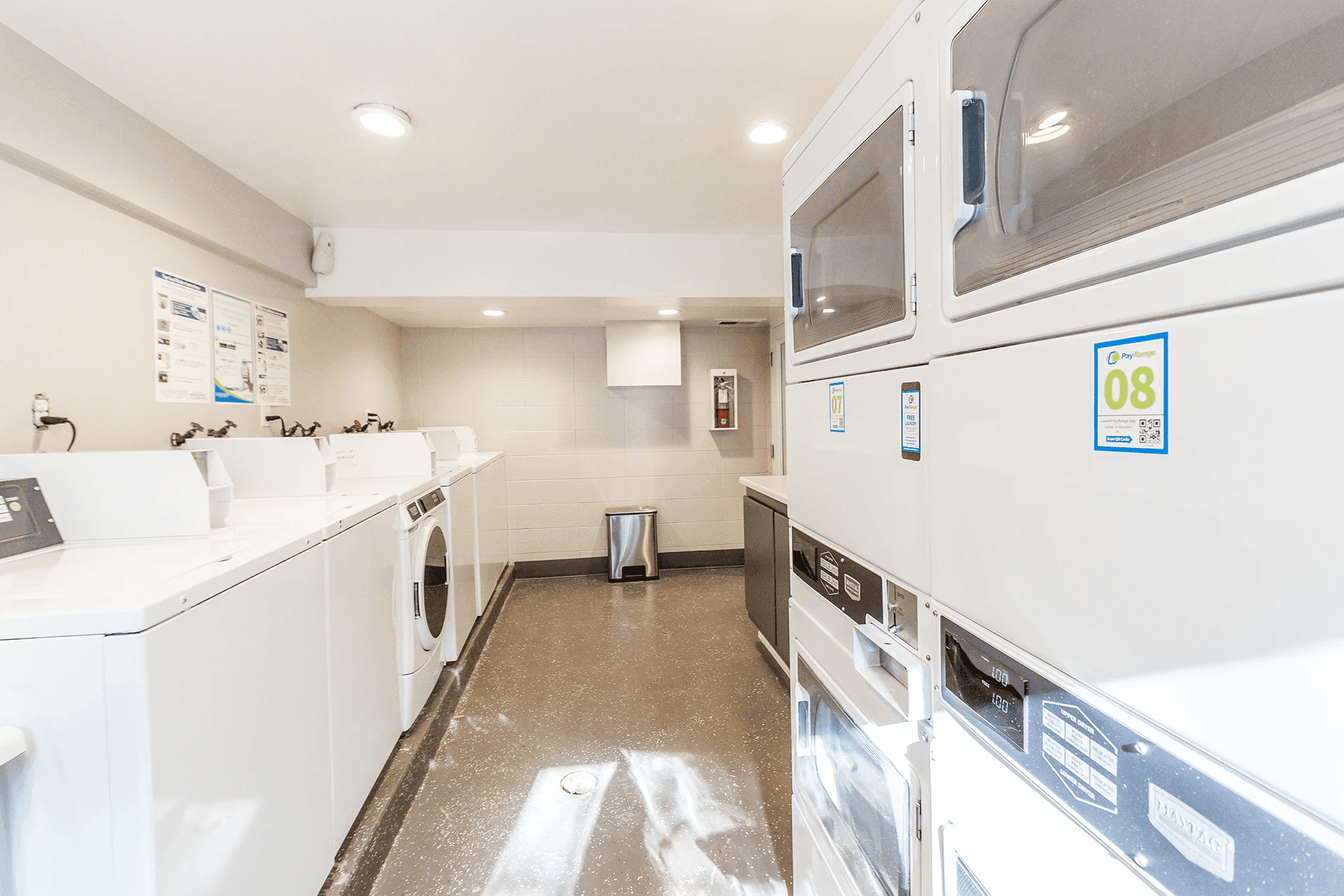 A bright, clean laundry room on Campus Way in Provo, featuring several white washing machines and dryers lined along the walls, a trash can in the corner, and informational posters decorating the space.