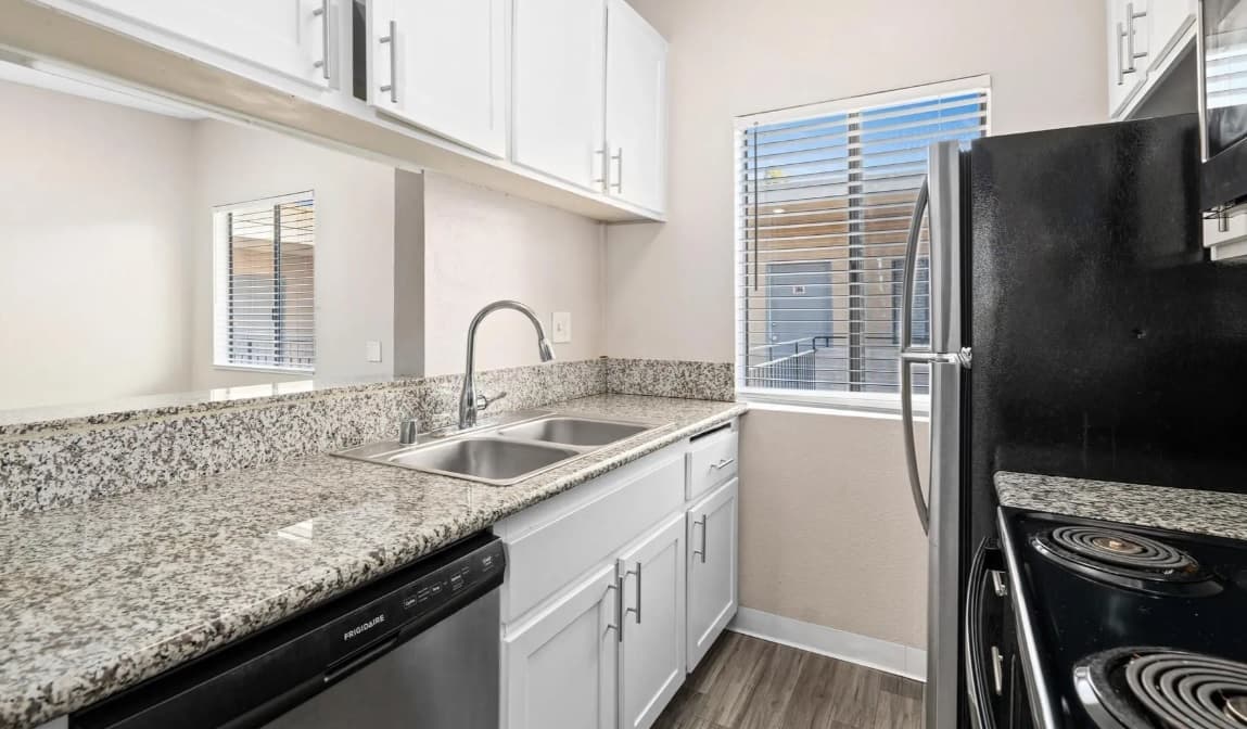 A modern kitchen at Campus Village Apartments features granite countertops, white cabinets, a stainless steel dishwasher, double sink, black stove and refrigerator, window with blinds, and wood-style flooring.