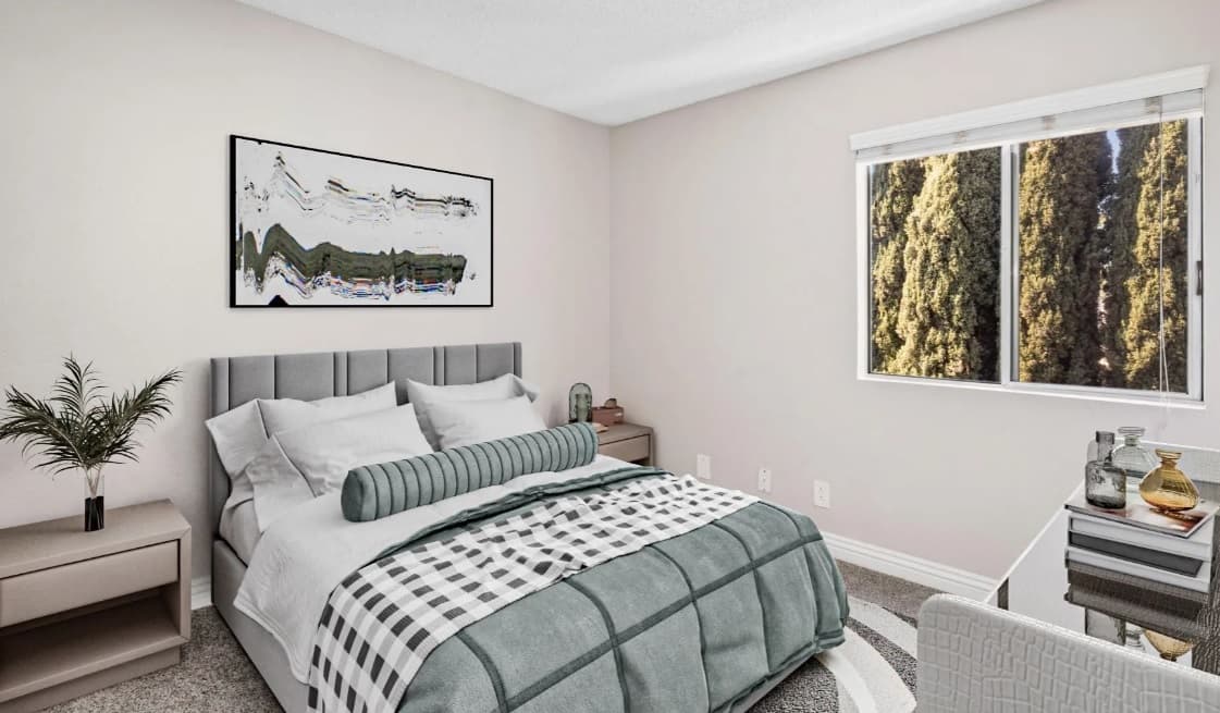 A modern bedroom at Campus Village Apartments features a gray upholstered bed, white and gray bedding, two nightstands, a large abstract painting above the bed, a potted plant, a window with views of tall trees, and decorative vases on the dresser.
