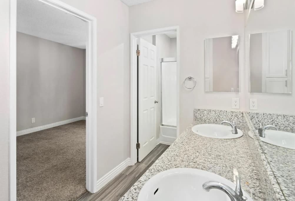 A bathroom at Campus Village Apartments features a granite countertop with two sinks, a large mirror, white cabinets, and a door leading to a carpeted bedroom. A shower with a white curtain is visible through another open door.