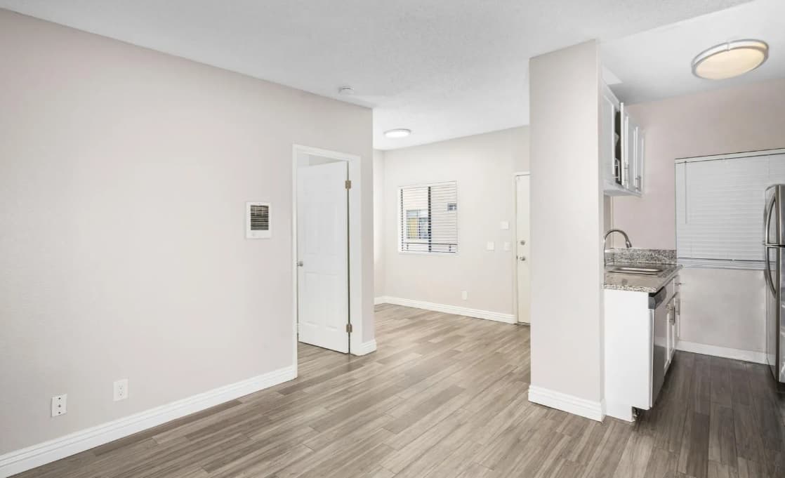 A small, modern Campus Village Apartments interior with light gray walls, wood-style flooring, a white door, and a compact kitchen featuring granite countertops and stainless steel appliances. Two windows provide natural light.