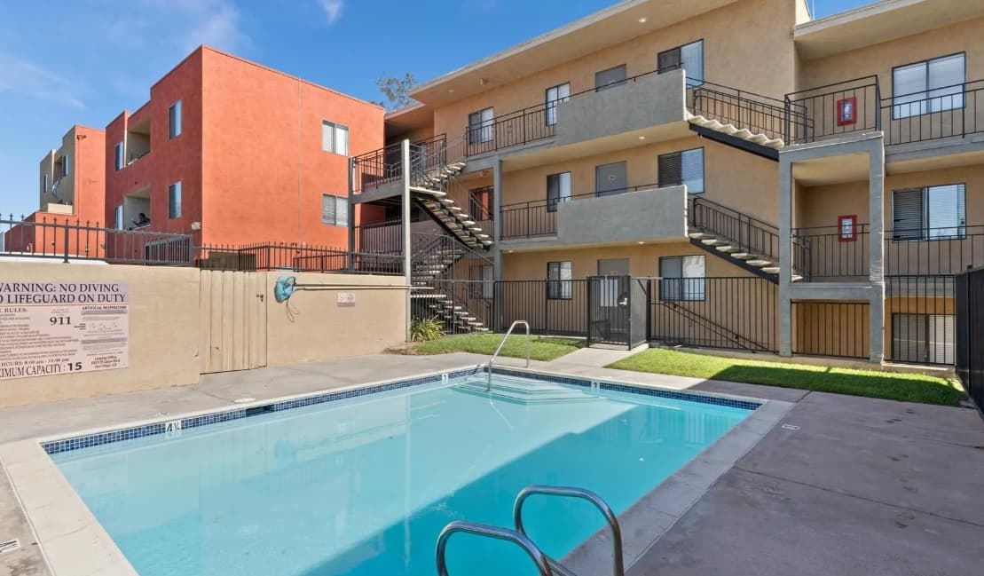 A small outdoor swimming pool sits in front of the Campus Village Apartments, a two-story building with stairs and balconies. The fenced area features pool rules on the wall and a life ring hanging nearby for safety.