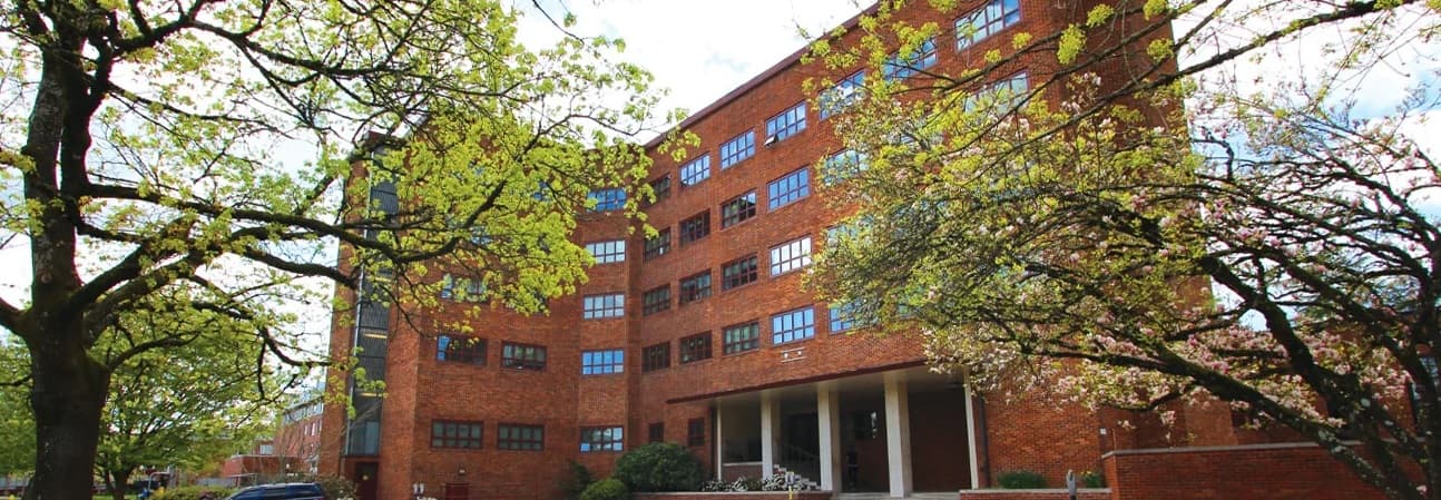 A large red-brick building with multiple rows of windows and a main entrance with columns, surrounded by leafy green trees on a bright, partly cloudy day.