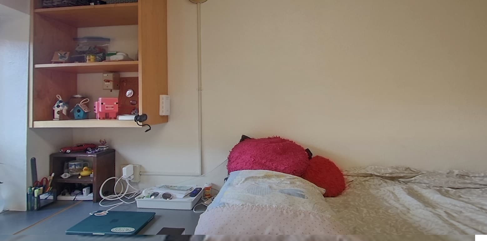 A cozy bedroom with a neatly made bed, a pink plush cat pillow, wall shelves holding books, toys, and containers, and a small desk below with stationery, cords, and electronic devices against a plain wall.
