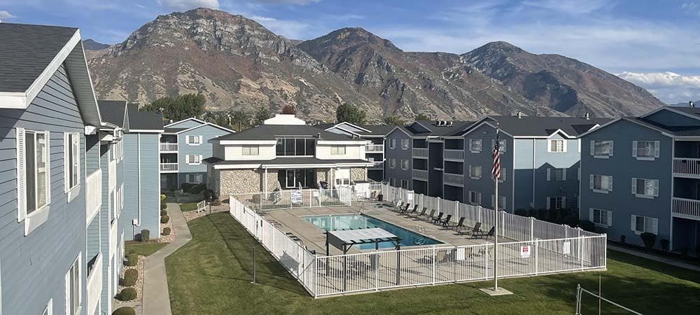 An outdoor swimming pool at Carriage Cove Provo is surrounded by a white fence, nestled between light blue apartment buildings, with mountains in the background under a partly cloudy sky.