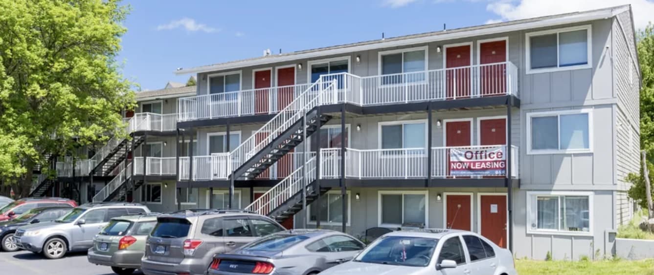 A three-story apartment building, College Crest, features red doors, exterior stairs, parked cars in front, and a sign reading Office Now Leasing on the railing. Trees are visible on the left side.
