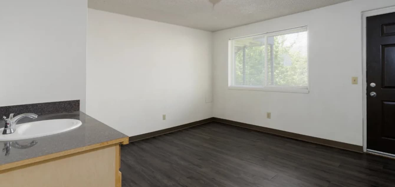 A small, empty room at College Crest with dark wood flooring, white walls, a window with blinds letting in natural light, a black door, and a corner countertop with a sink visible on the left.