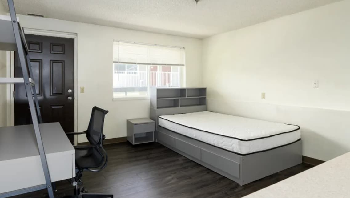 A minimalist bedroom at College Crest with a gray twin bed, a small nightstand, a desk with a black office chair, and a window with blinds. The flooring is dark wood, and the walls are plain white.