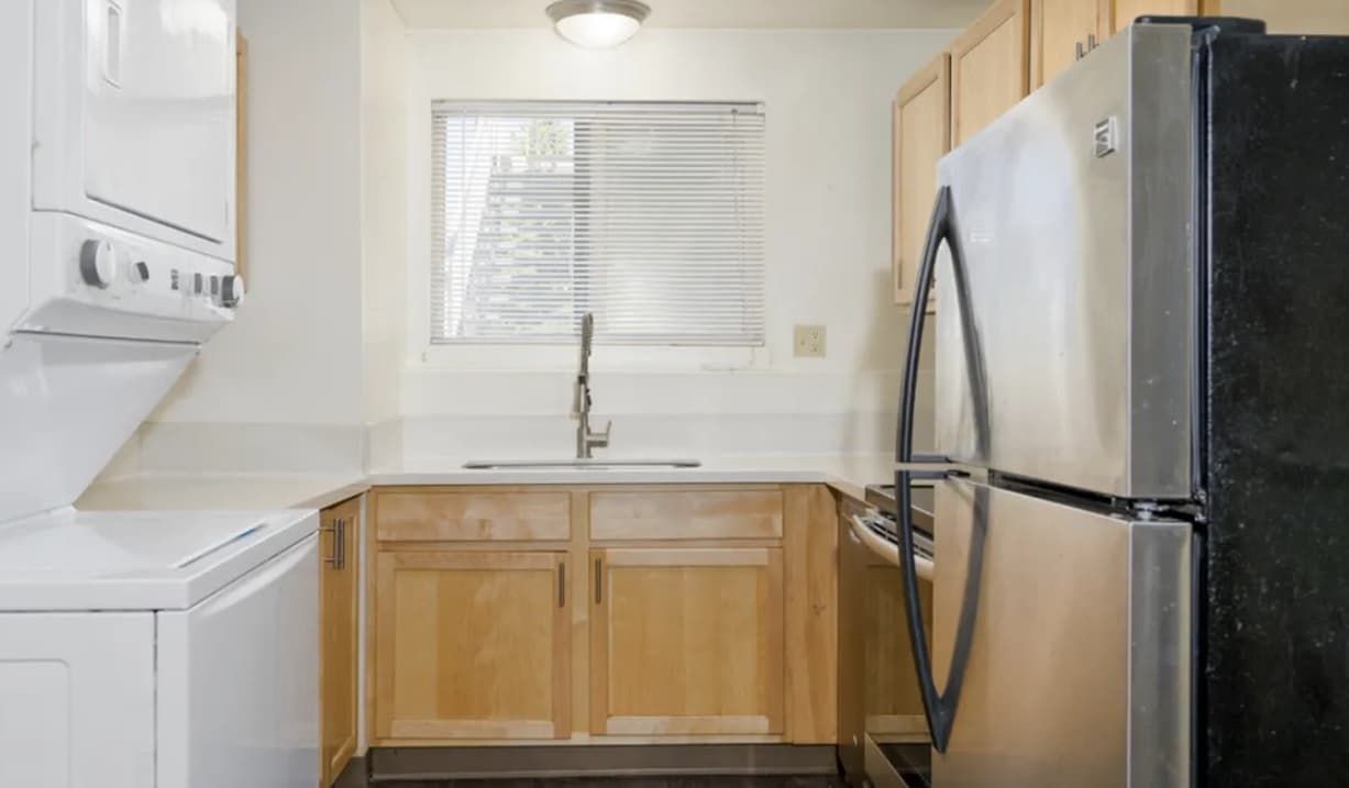A compact College Crest kitchen with light wood cabinets, a stainless steel refrigerator, a built-in oven, a sink under a window with blinds, and a stacked washer and dryer unit to the left.