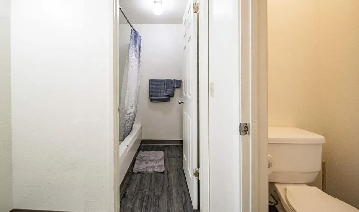 A bathroom at College Crest features gray wood-style flooring, a white bathtub with a blue and white shower curtain, gray towels on a rack, a white toilet, and a small gray bath mat on the floor.