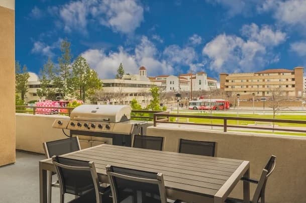 Outdoor patio with a dining table, chairs, and a barbecue grill at Cerca Student Housing, overlooking a campus with trees, buildings, and a red bus under a bright blue sky with scattered clouds.