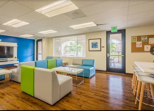 A modern lounge at Cerca Student Housing with colorful sofas, white tables, a TV, wall art, wood flooring, and windows letting in natural light. High chairs line a counter near a glass door leading outside.