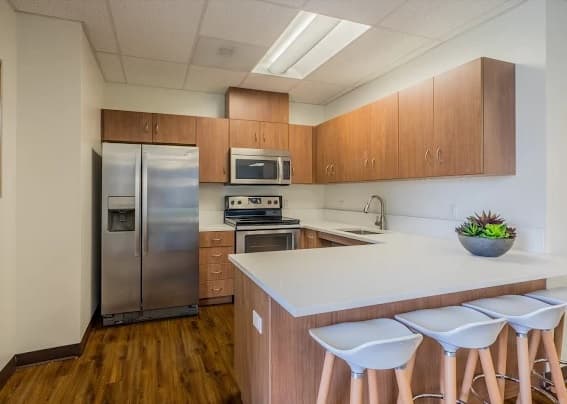 Modern kitchen at Cerca Student Housing with wooden cabinets, stainless steel appliances, white countertops, built-in oven, and refrigerator. Three white barstools line the island, topped with a bowl of succulents.