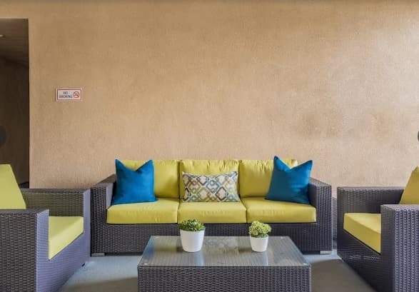Outdoor seating area at Cerca Student Housing features a yellow-cushioned sofa, matching armchairs, blue and patterned pillows, a glass coffee table, and potted plants. A No smoking sign is visible on the beige wall.