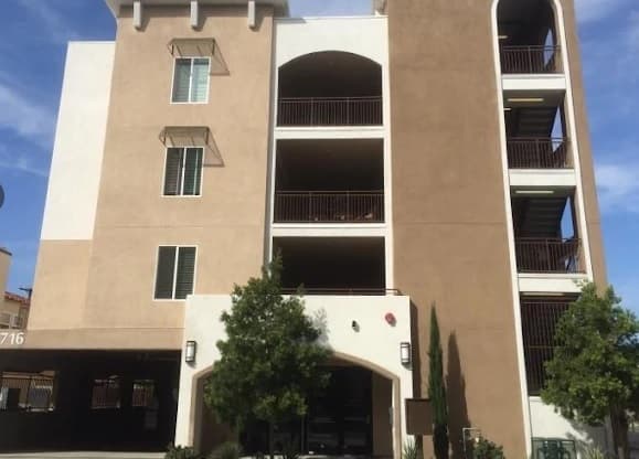 A four-story modern apartment building, ideal for Cerca Student Housing, features tan and white exterior walls, balconies with dark railings, a ground-level parking area, and two trees planted in front of the entrance.