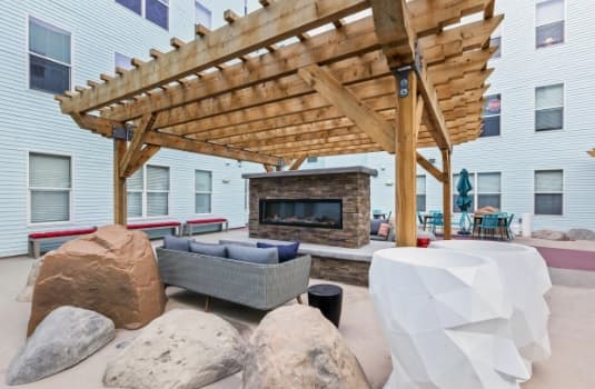 Outdoor courtyard at Canyon Flats Reno features a wooden pergola, stone fireplace, gray sofa, large decorative rocks, white geometric sculpture, and patio seating, all surrounded by a light blue building with multiple windows.