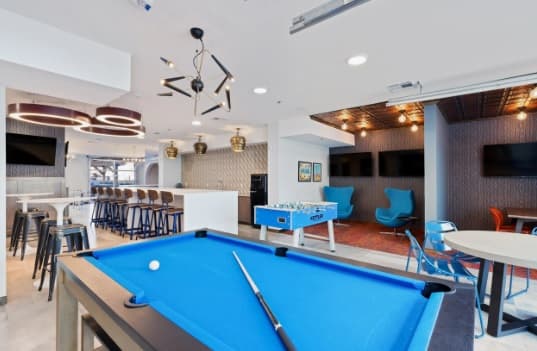 Modern game room at Canyon Flats Reno with a blue pool table in the foreground, foosball table, several tables and chairs, bar seating, large TVs on the walls, and contemporary lighting fixtures in a bright, open space.