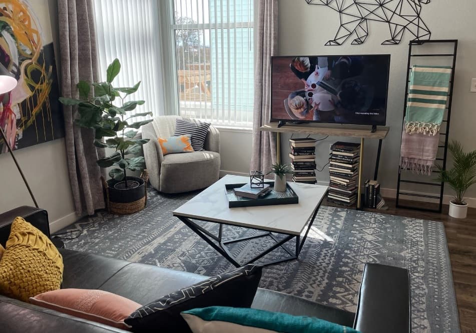 A modern living room at Canyon Flats Reno features a gray sofa, patterned rug, TV on a stand, stacks of books, a large plant, artwork on the walls, and sunlight streaming through tall windows.
