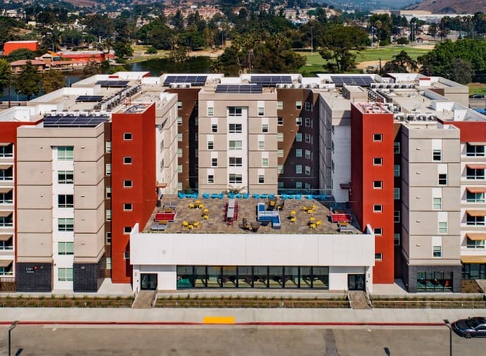 Aerial view of a modern multi-story apartment building with red accents, a large rooftop patio with colorful seating, and solar panels on the roof. Trees, hills, and a golf course are visible in the background.