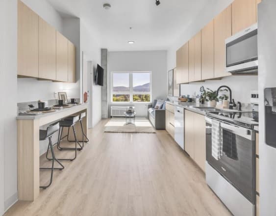 Modern apartment kitchen with light wood cabinets, stainless steel appliances, a small dining area for two, and a bright living space with mountain views—perfect for student housing at Chapter at Eugene.