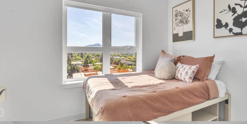 A neatly made bed with brown and white bedding sits by a large window at Chapter at Eugene, offering city and mountain views. Two framed botanical prints hang on the white wall above the bed.