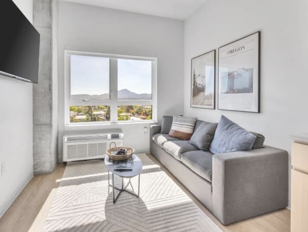Bright modern living room at Chapter at Eugene with large windows, gray sofa, round coffee table, wall-mounted TV, framed posters, and light wood flooring. Sunlight fills the space, creating a cozy atmosphere.
