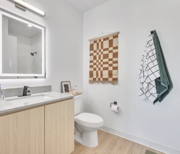 Modern bathroom at Chapter at Eugene with a light wood vanity, a large LED-lit mirror, a toilet, a brown and white patterned wall hanging, and two towels—one green and one white with black grid lines—on the wall.