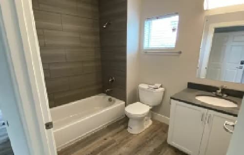 Modern bathroom with a bathtub and shower on the left, a toilet in the center, and a white vanity with a sink and mirror on the right. Wood-look flooring and a small window above the toilet.