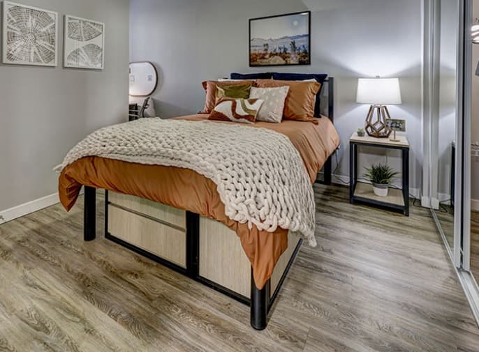A cozy bedroom with a neatly made bed featuring orange bedding and a chunky knit blanket, wooden floors, a bedside table with a lamp and plant, and wall art above the bed. Storage bins are under the bed.