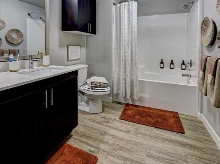Modern bathroom with wood-look flooring, dark cabinets, a white countertop, two rust-colored rugs, a bathtub with a white curtain, and decorative wall plates and towels in warm neutral tones.