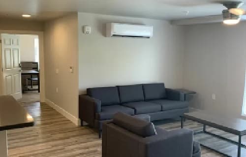 A modern living room at Coffee House Apartments features light-colored walls, wood-style flooring, a dark gray sofa set, a coffee table, and a wall-mounted air conditioning unit. An open doorway leads to another room with a desk and chair.