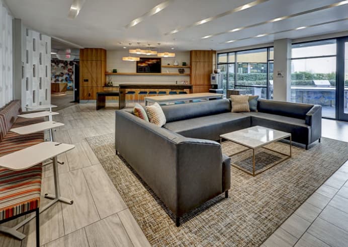 Modern lounge area with a large gray sectional sofa, a marble coffee table on a patterned rug, striped bench seating with white side tables, and a communal wooden table in a bright room with large windows.