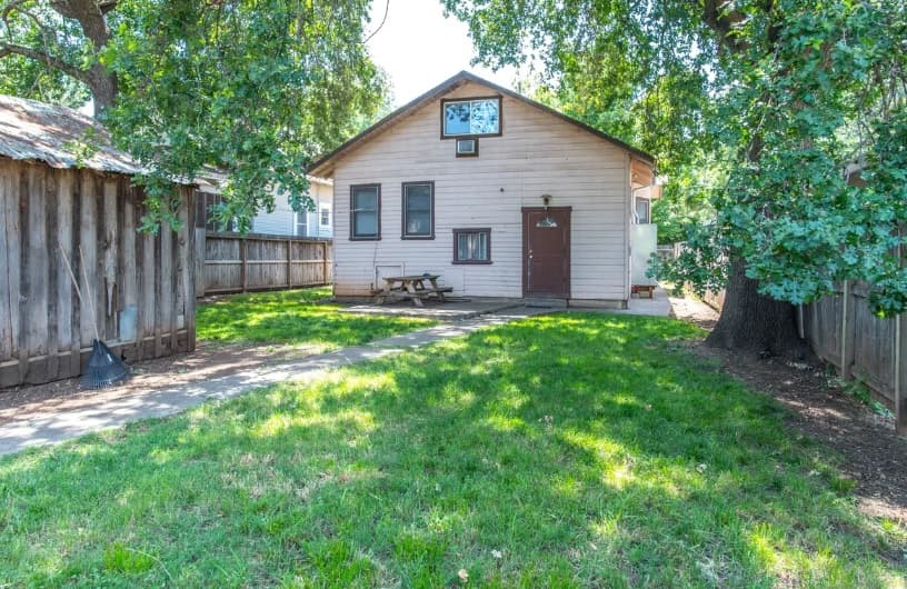 A small beige house with a brown door and windows sits in a grassy backyard—perfect for College Housing Chico. There’s a wooden picnic table near the door, a large tree on the right, a wooden shed on the left, and a path leading to the house.