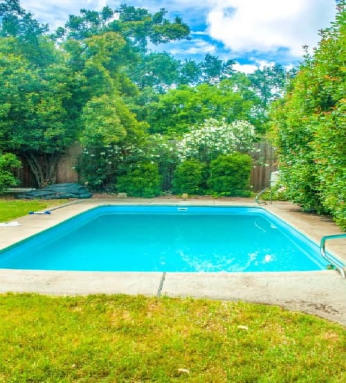 A clean, rectangular swimming pool is surrounded by grass, bushes, and trees in a lush backyard under a partly cloudy sky—an inviting retreat perfect for College Housing Chico students. Steps and a metal handrail are visible at one end of the pool.