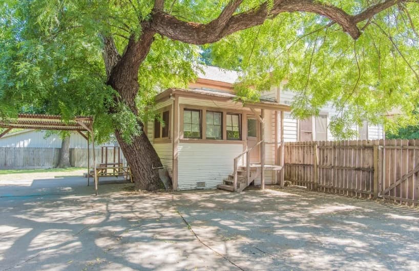A small white house, perfect for College Housing in Chico, sits under a large, leafy tree. A wooden fence and a carport are on either side, with sunlight filtering through branches and casting shadows on the driveway.