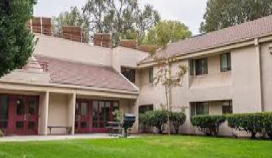A beige two-story building with red doors and windows, surrounded by trimmed bushes and green grass—ideal Chico Student Housing; tall trees are visible in the background under a cloudy sky.