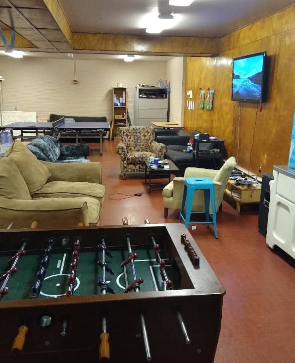 A cozy iHouse game room in Corvallis with a foosball table in the foreground, comfy couches and chairs, a TV on the wall showing a video game, plus a ping pong table and shelves in the background.