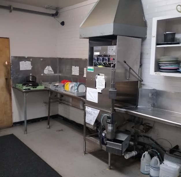 A commercial kitchen dishwashing area at iHouse in Corvallis features a large industrial dishwasher, clean and dirty dish racks, a sink, and cleaning supplies under the counter. White tiled walls and shelves hold plates and bowls.