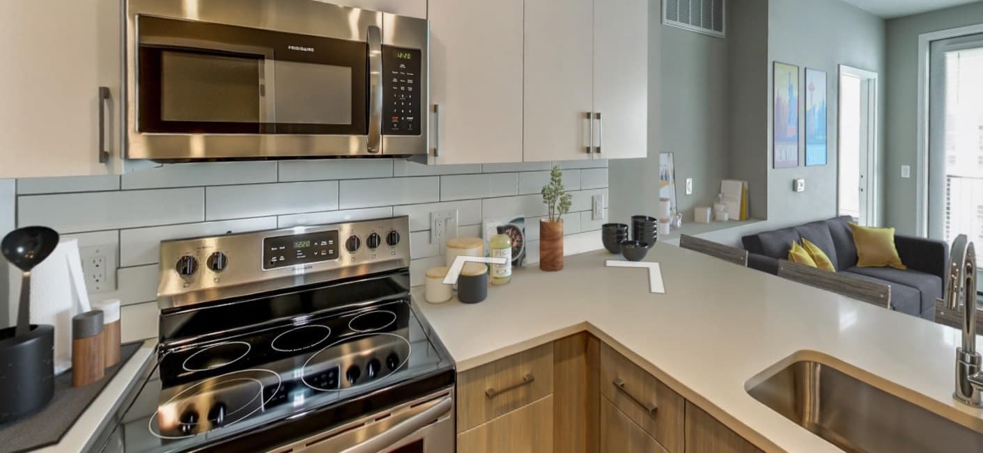 Modern kitchen with stainless steel appliances, white backsplash, and wood cabinetry. The countertop features kitchen items and a sink. In the background, a living room with a gray sofa offers comfort—experience elevated living at CoLab Denver.